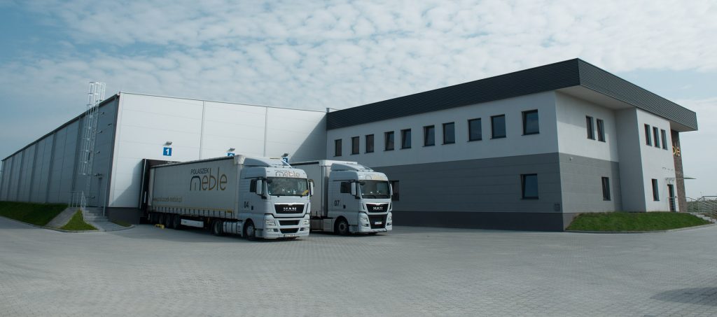 Polaszek Meble Dwie ciężarówki dostawcze zaparkowane przed nowoczesnym budynkiem magazynu przemysłowego z widocznym oznakowaniem Meble Chojnice. Chojnice