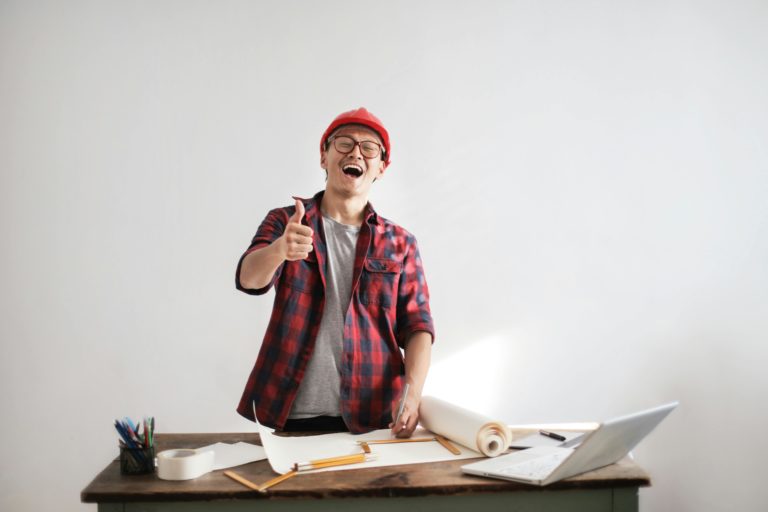 Polaszek Meble Wesoła osoba w czerwonym kapeluszu i koszuli w kratę stoi przy biurku z materiałami projektowymi i laptopem, pokazując kciuki do góry. Chojnice