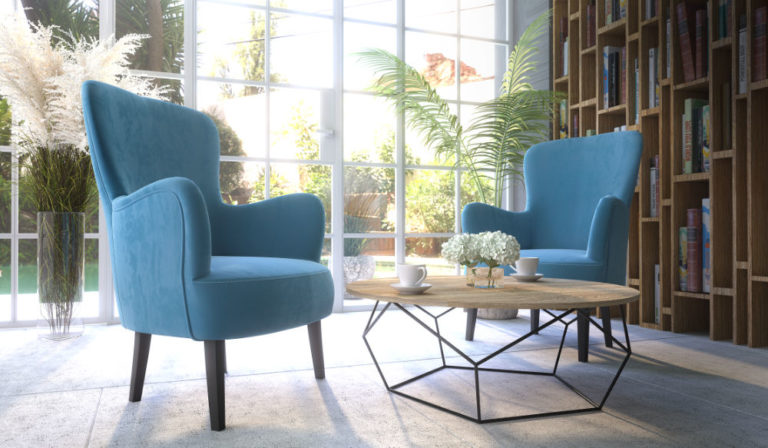 Polaszek Meble Dobrze oświetlony pokój z dwoma niebieskimi fotelami, drewnianym stolikiem kawowym z białymi kwiatami i dwiema filiżankami. W tle znajdują się duże okna, rośliny i półki na książki. Chojnice