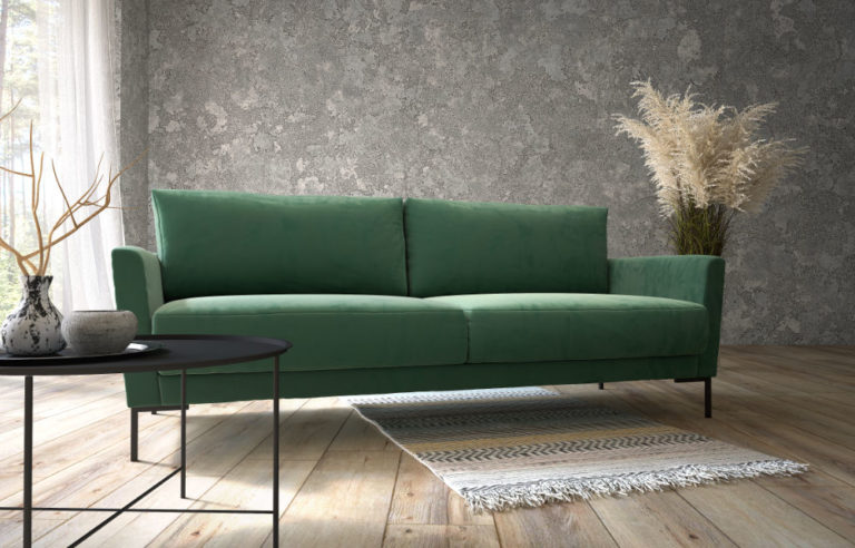 Polaszek Meble Zielona sofa stoi na tle szarej ściany z teksturą, z okrągłym czarnym stolikiem kawowym z przodu i wazonem z trawą pampasową po prawej stronie. Mały dywanik leży na drewnianej podłodze pod sofą. Chojnice