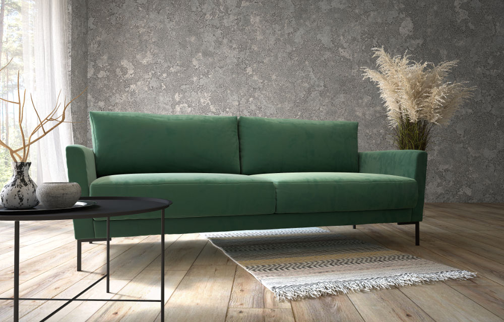 Polaszek Meble Zielona sofa stoi na tle szarej ściany z teksturą, z okrągłym czarnym stolikiem kawowym z przodu i wazonem z trawą pampasową po prawej stronie. Mały dywanik leży na drewnianej podłodze pod sofą. Chojnice